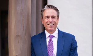 Portrait of Dr. Randall Blazic smiling, wearing a blue suit jacket, white shirt, and purple patterned tie, leaning against a wooden pillar